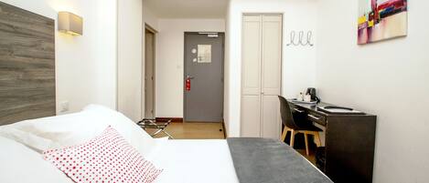 A hotel room with a bed, a desk, and a chair. There is a door leading to another room and a wall-mounted artwork.