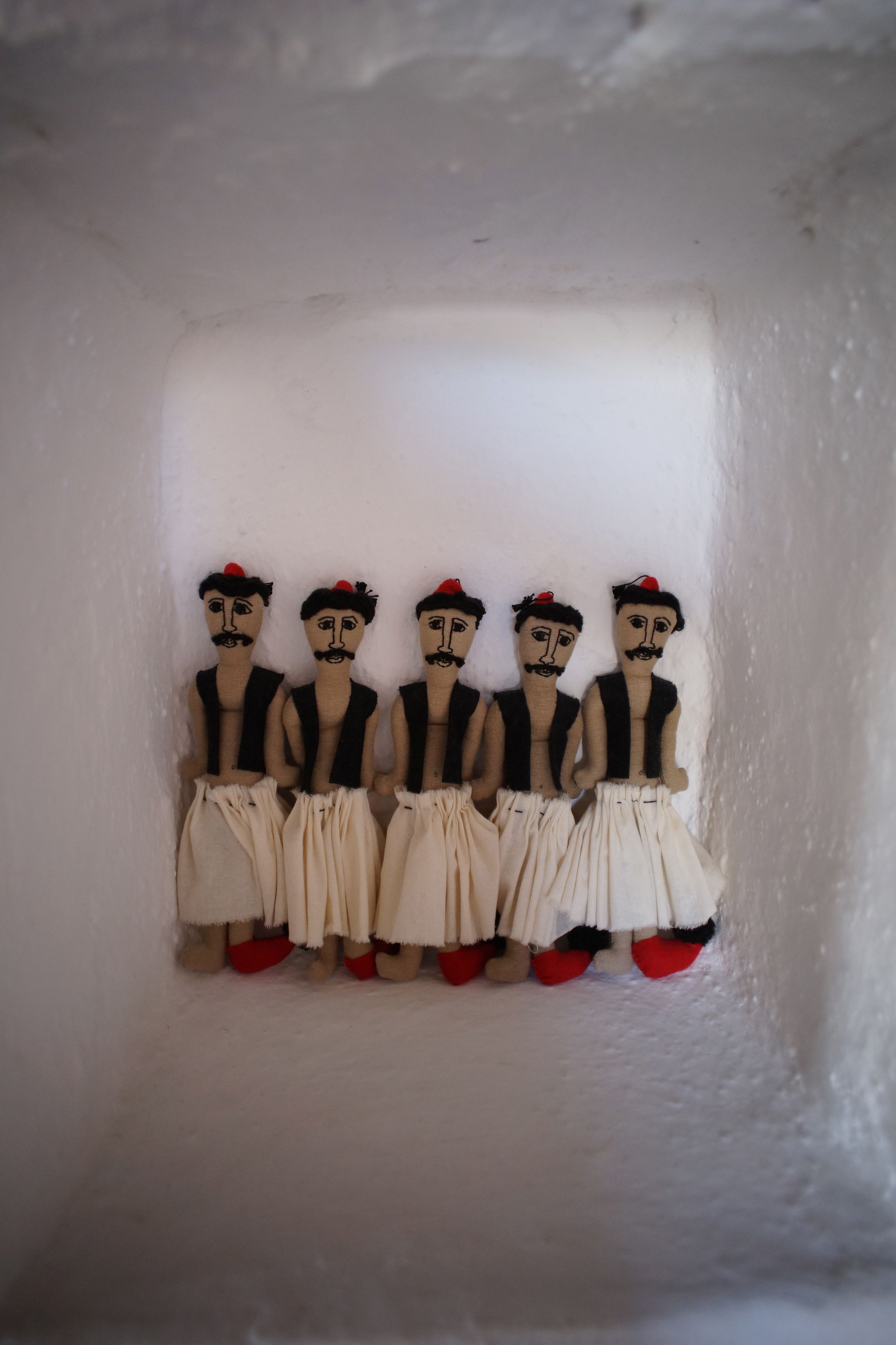 Five wooden figures with black and white clothing, red hats, and red shoes, standing in a row against a white wall.