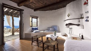 Um quarto com teto de madeira, parede branca, janela, um banco, uma mesa e vista para o mar.