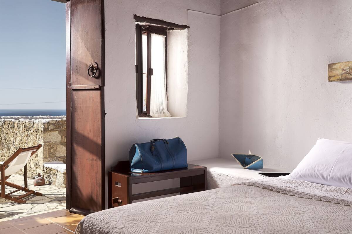 A bedroom with a bed, a wooden nightstand, a blue bag, a white pillow, and a view of the sea through a window.