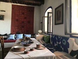 Un comedor con una mesa dispuesta para cuatro personas, un sofá con cojines estampados, un tapiz rojo y una ventana con vistas.