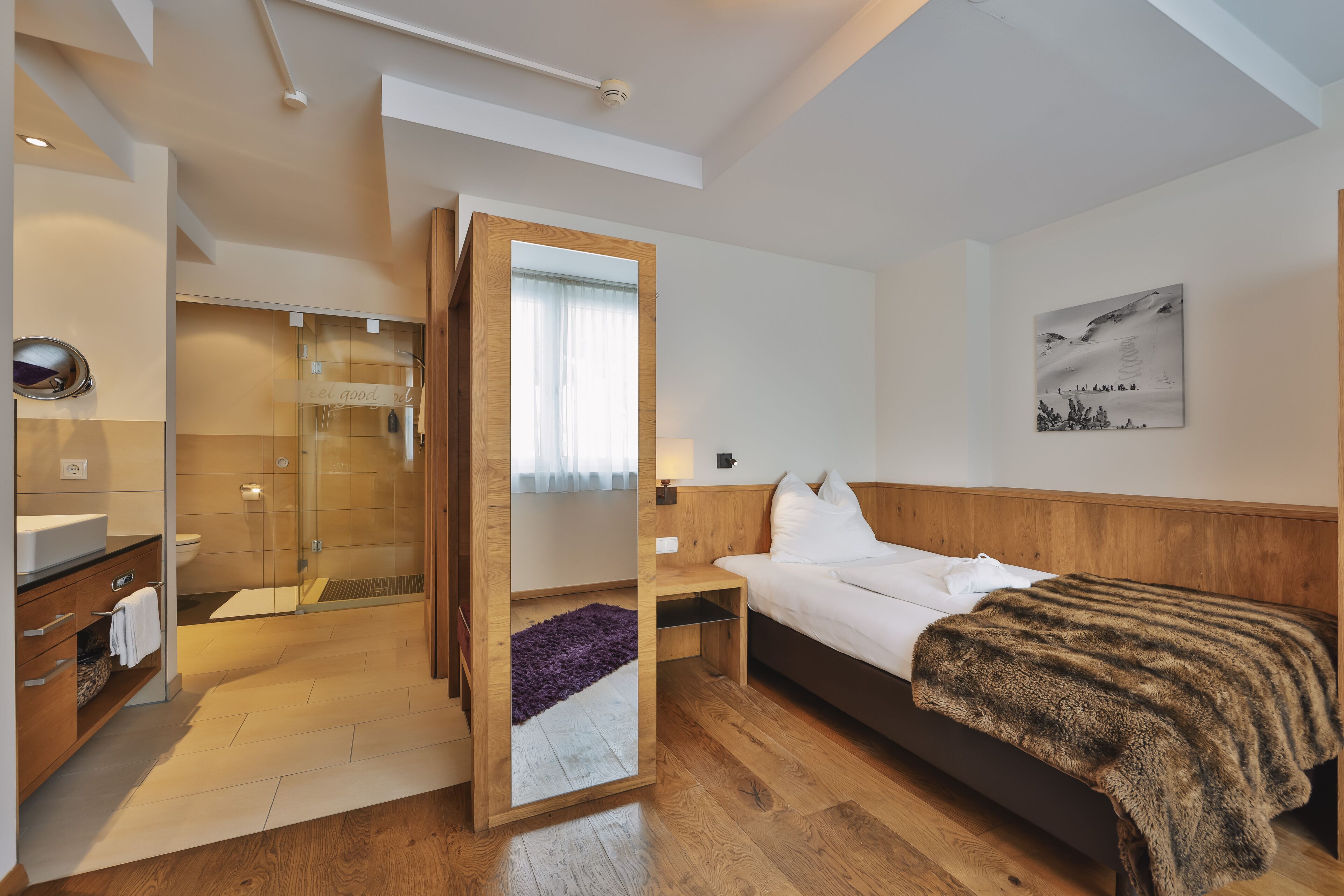 Une chambre d’hôtel moderne avec un lit en bois, une table de chevet, une commode, un miroir et une salle de bain dotée d’une douche en verre, ainsi qu’une œuvre d’art accrochée au mur.