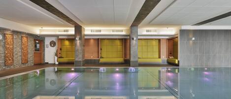 Una amplia piscina interior con agua cristalina, rodeada de zonas de estar con cojines amarillos y un diseño arquitectónico moderno.
