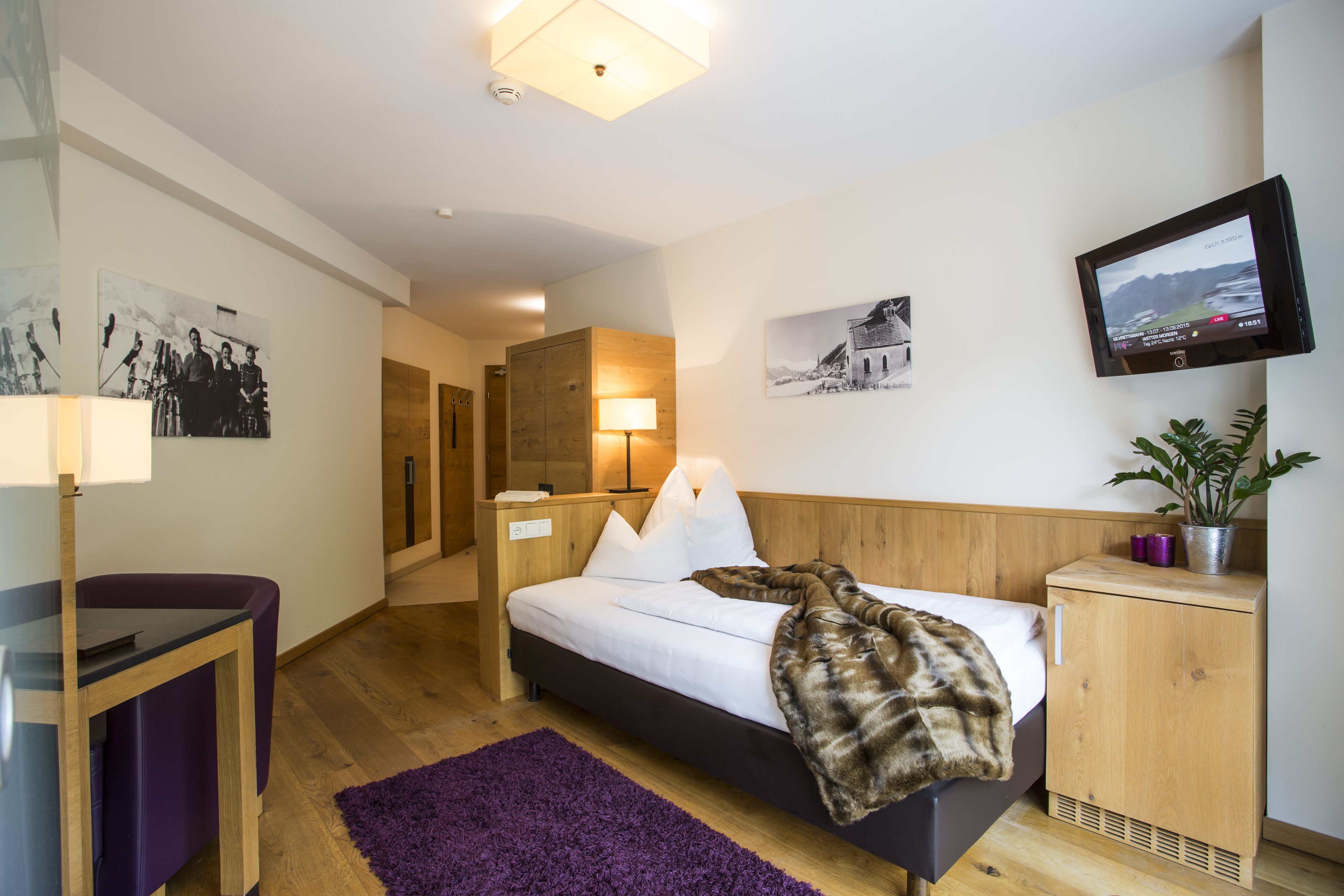 A modern hotel room with a bed, a desk, a TV, and artwork on the walls.
