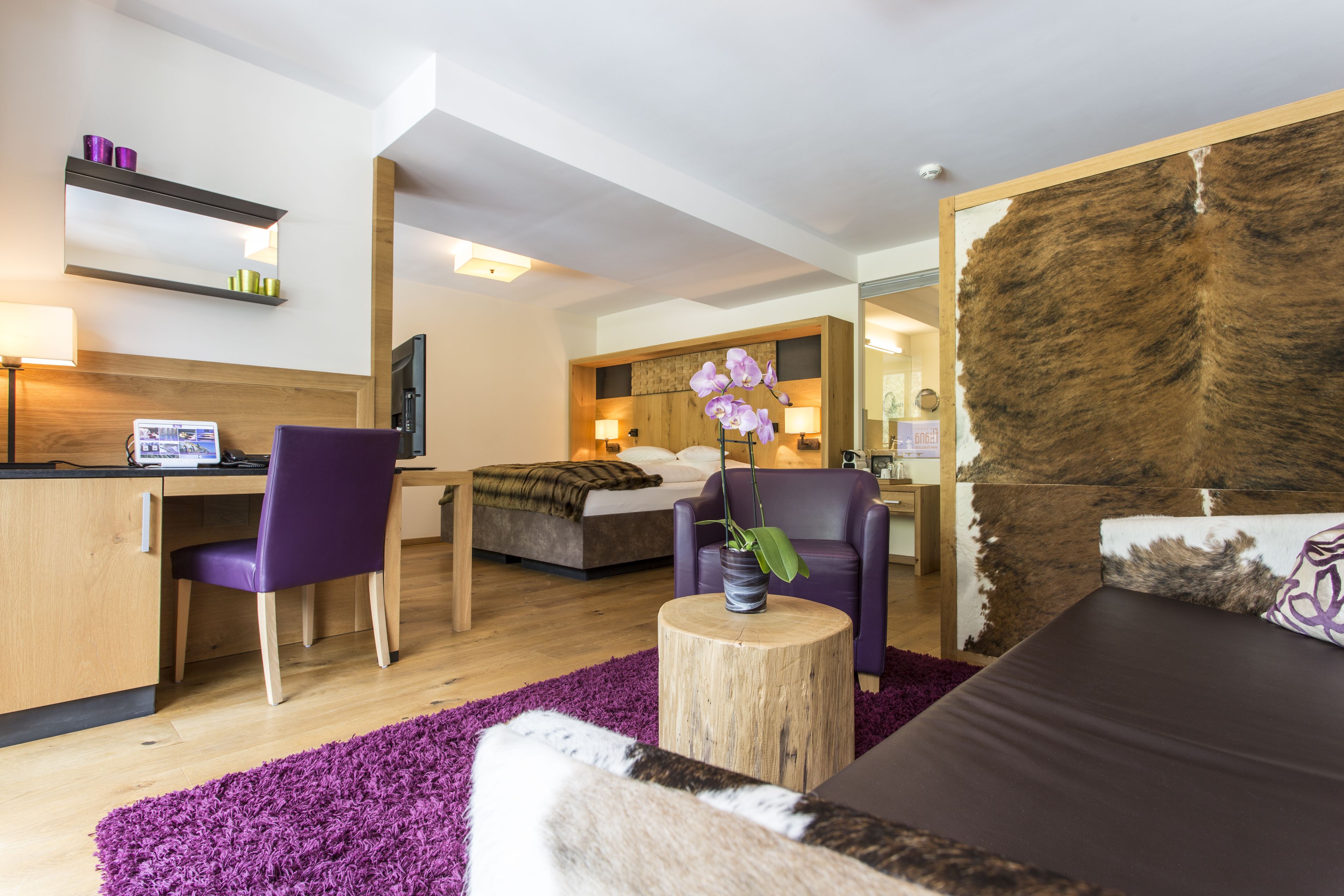 A modern hotel room with a wooden desk, purple chair, bed with a fur-like headboard, and a purple orchid plant.