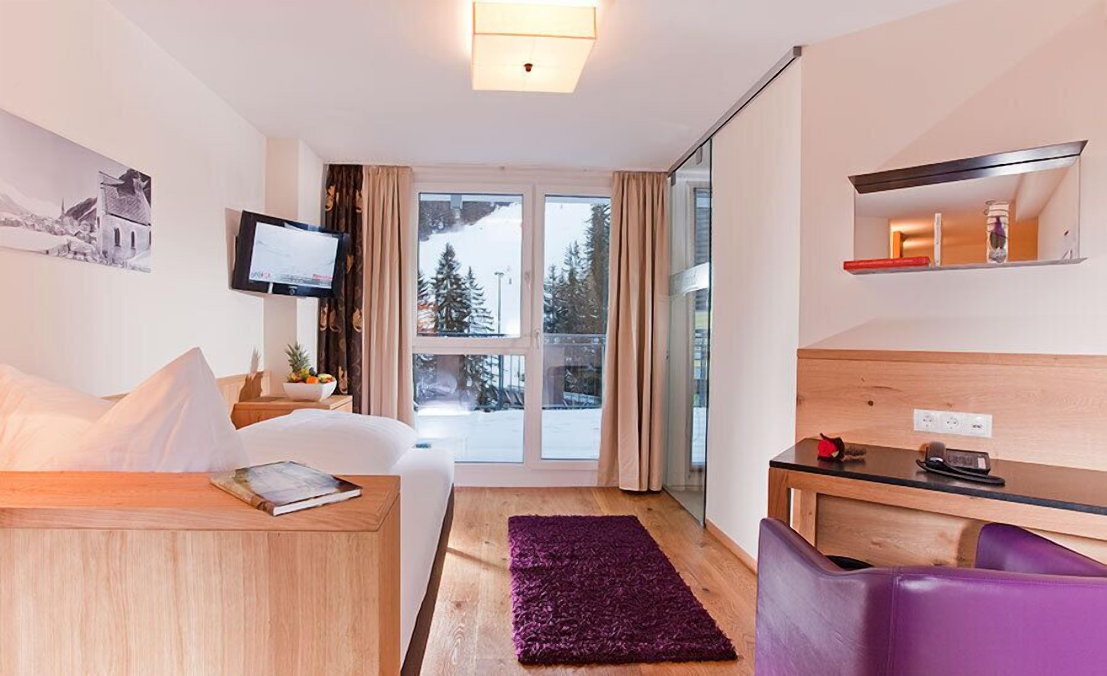 Hotelkamer met een bed, een bureau, een stoel, een televisie en een raam met uitzicht op bomen en een besneeuwd landschap.