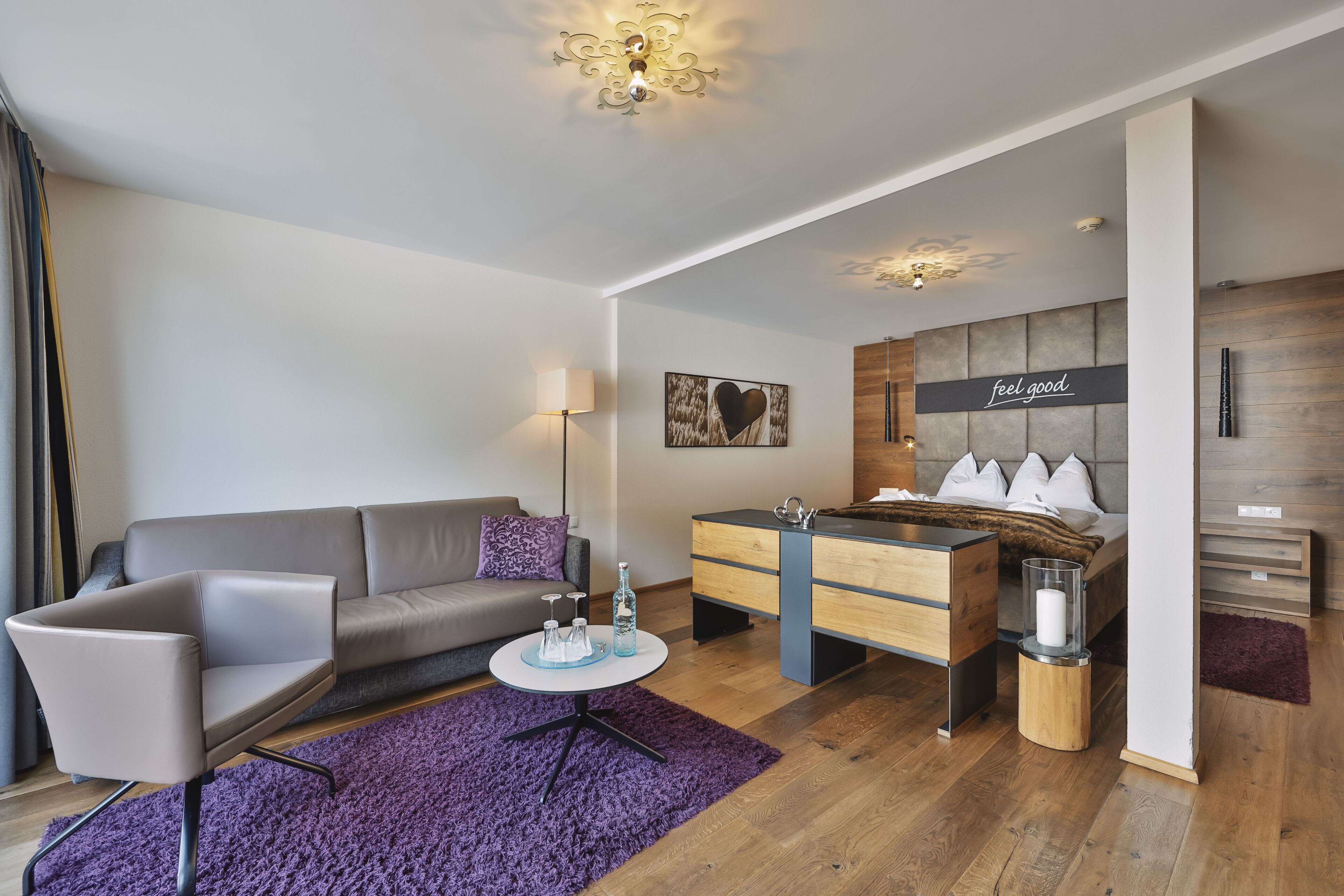 Una habitación de hotel moderna con un sofá gris, una pequeña mesa redonda, un aparador de madera y una cama con un cabecero que dice 'feel good'.