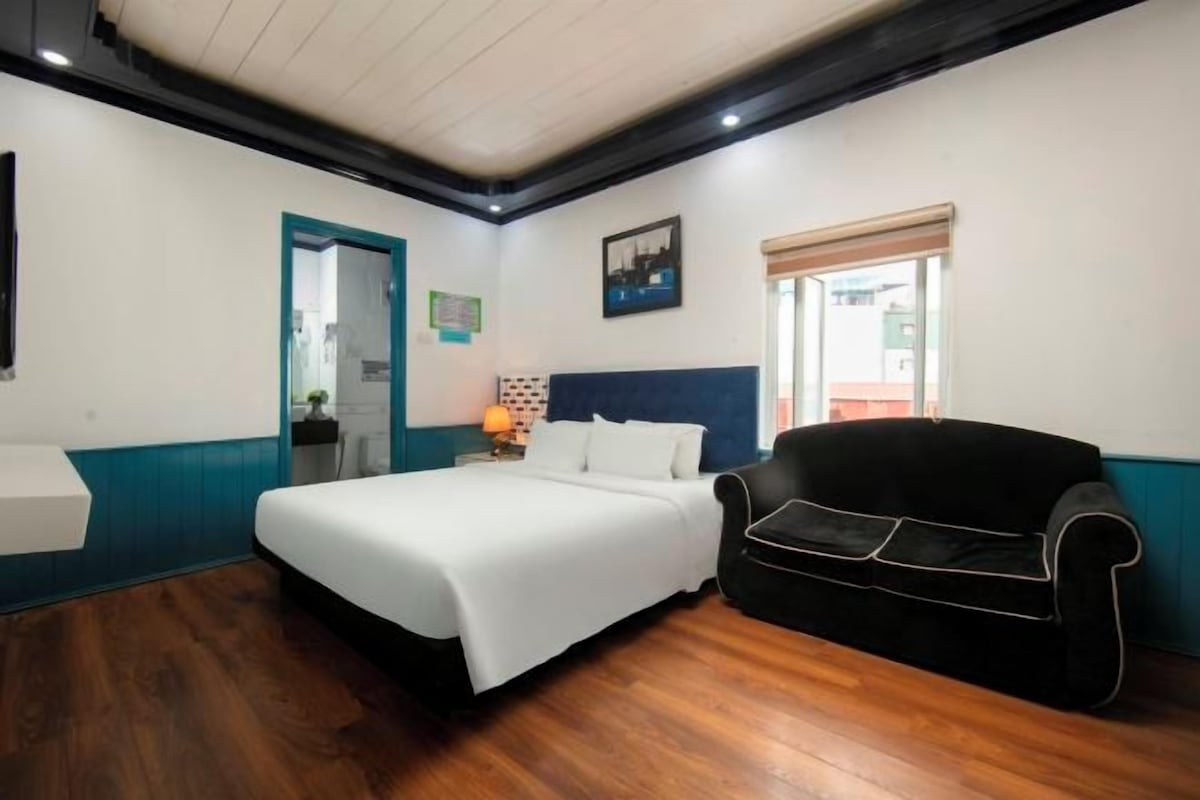 A hotel room with a bed, a black sofa, a wooden floor, a white wall, a blue accent wall, a window with blinds, and a picture on the wall.