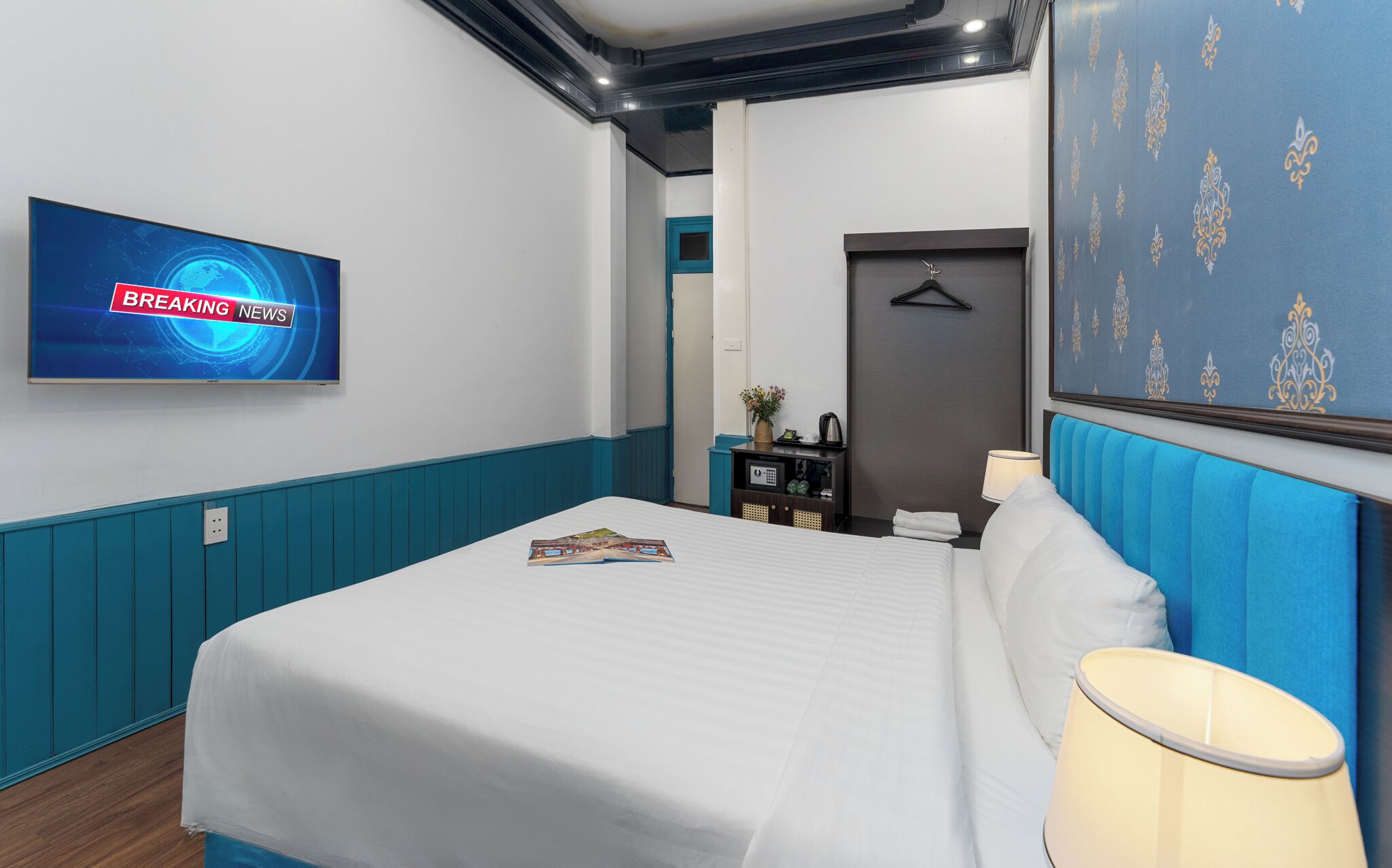 Habitación de hotel con cama, mesita de noche con lámpara, televisor mostrando 'BREAKING NEWS' y pared con diseño azul.