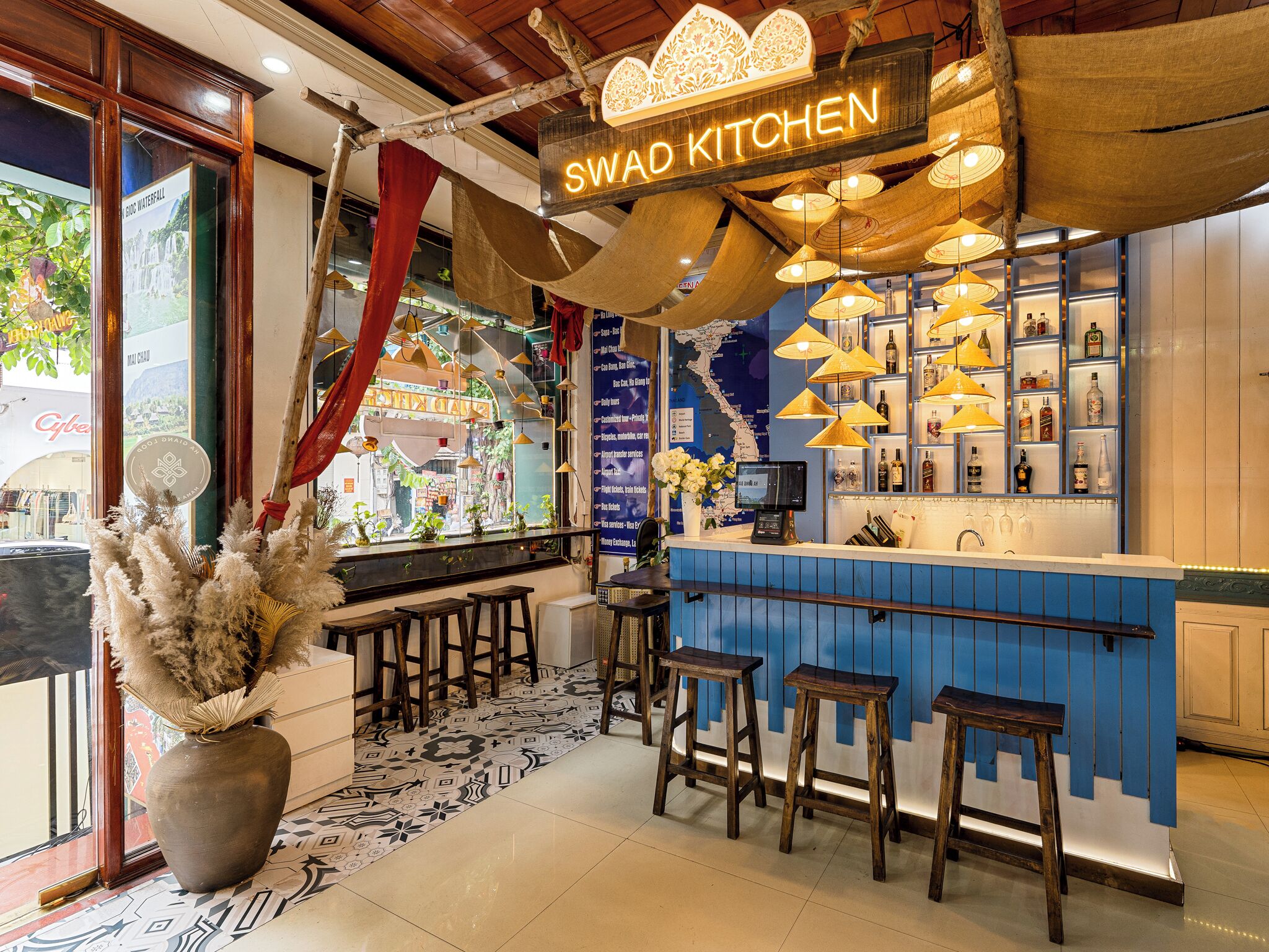 L’intérieur d’un restaurant avec un bar, des tabourets en bois, des luminaires suspendus et une enseigne indiquant « SWAD KITCHEN ».