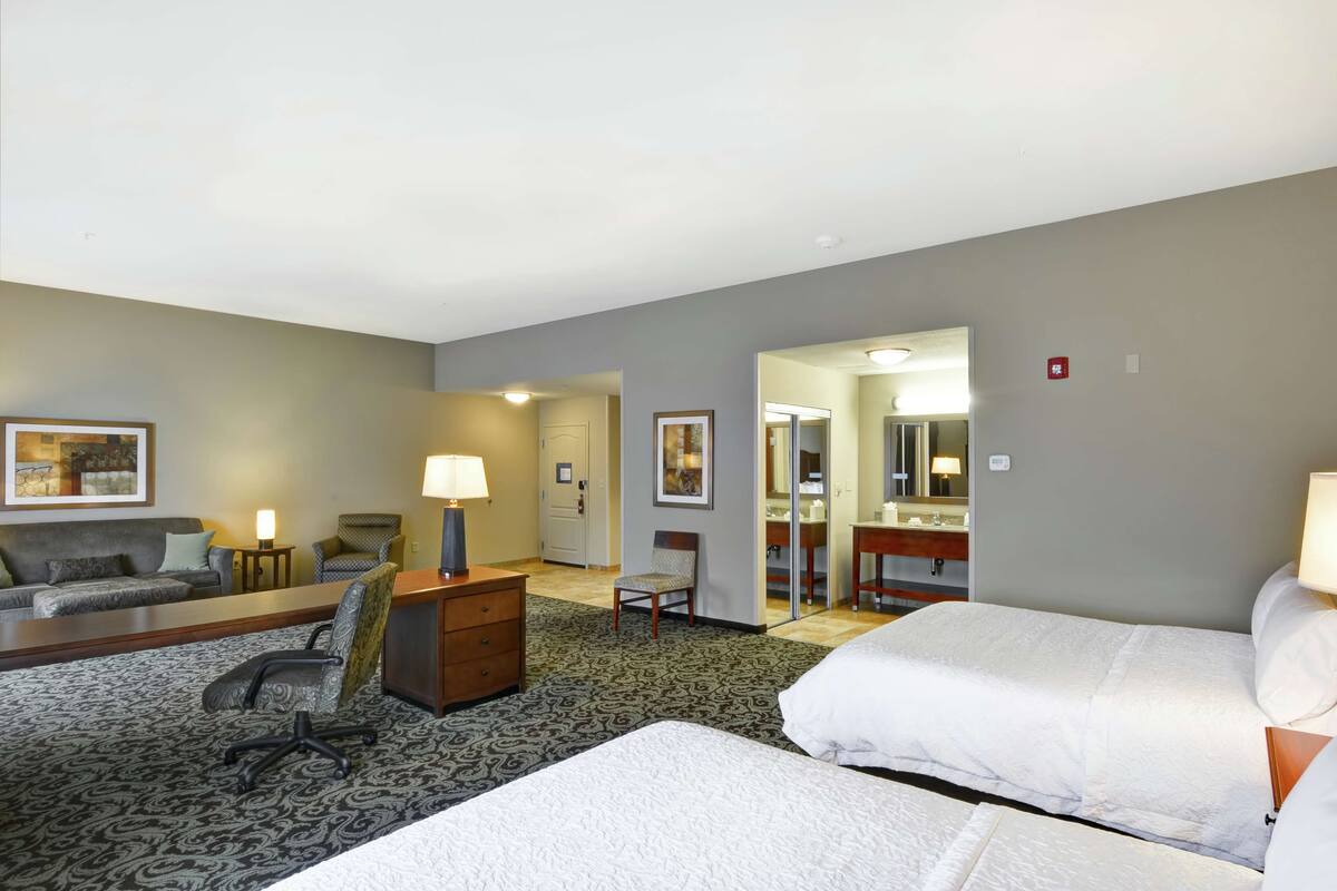 A hotel room with two beds, a desk, a chair, a lamp, a mirror, and a bathroom visible through an open door.