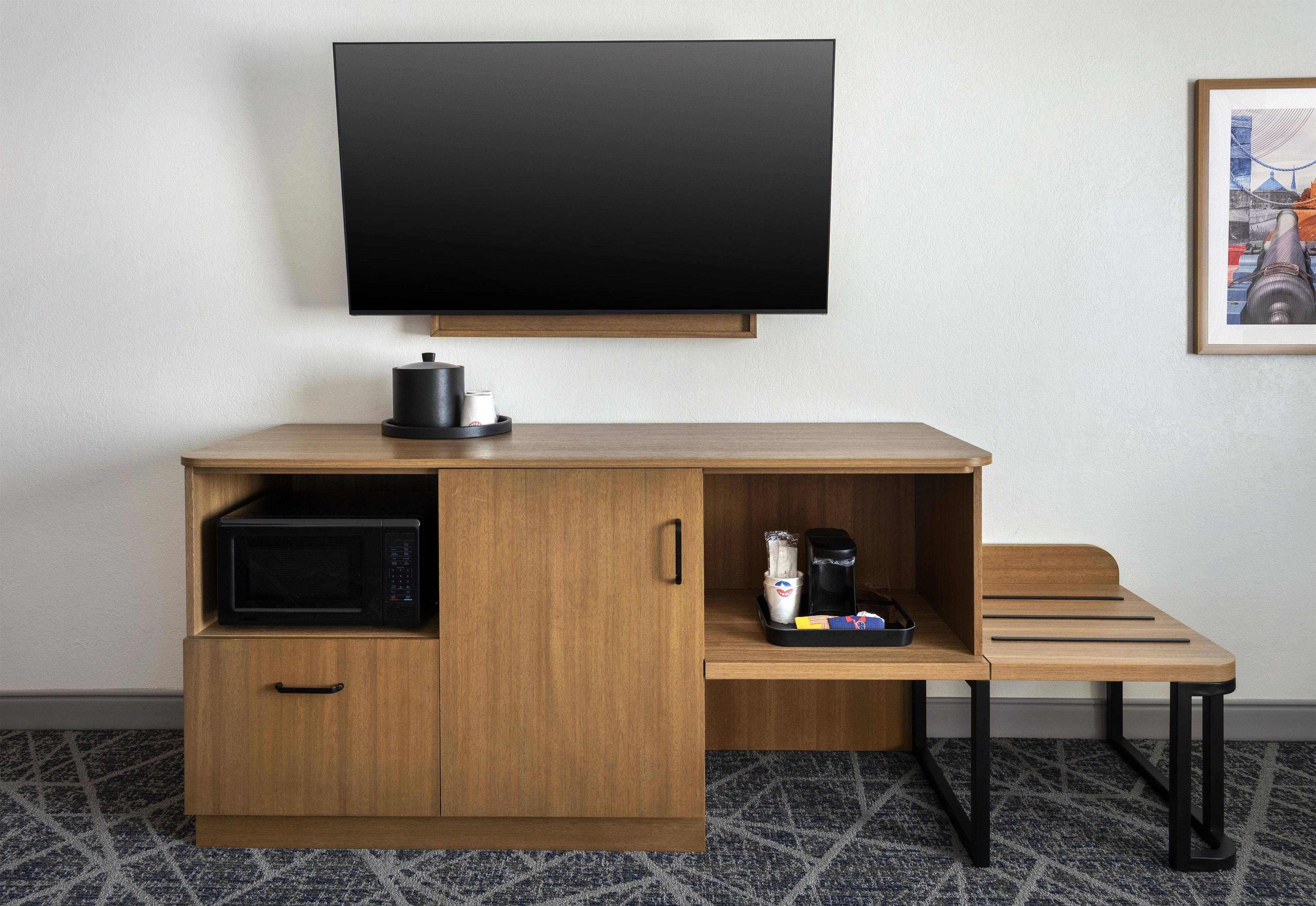 A compact hotel room with a wooden desk, a flat-screen TV, a microwave, and a coffee maker.