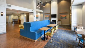 A hotel lobby with a blue sofa, wooden tables, and chairs. There are paintings on the walls and a TV mounted above a fireplace. - Hampton Inn and Suites New Hartford/Utica (New Hartford)