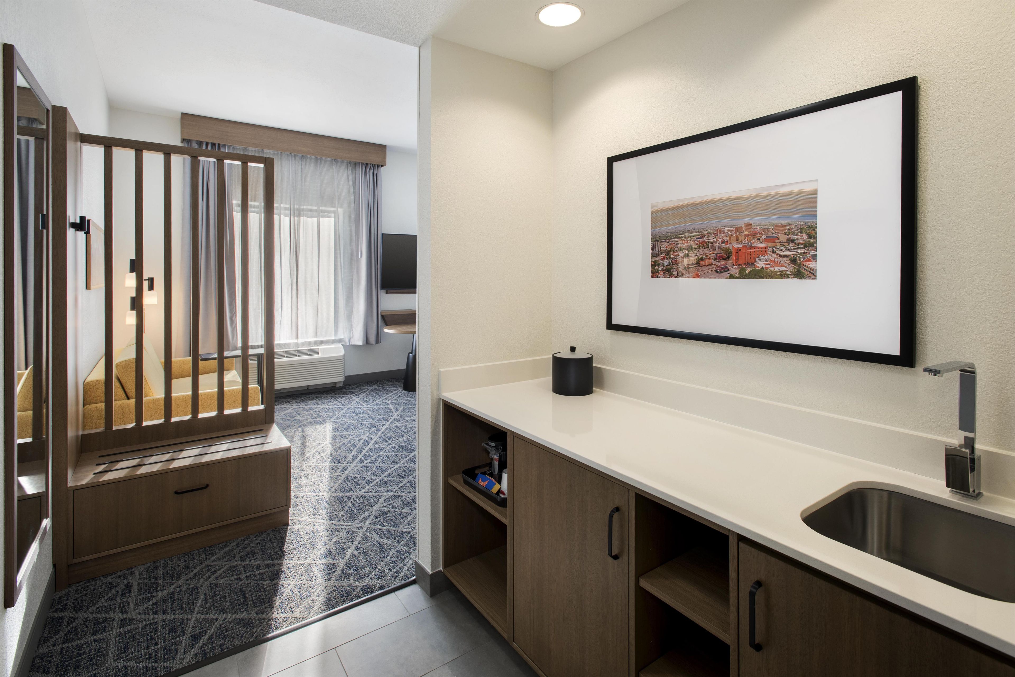 Une chambre d’hôtel moderne, équipée d’un lit, d’une salle de bain avec un lavabo et un miroir, d’un tableau encadré représentant un paysage urbain accroché au mur, et d’une fenêtre avec des rideaux.