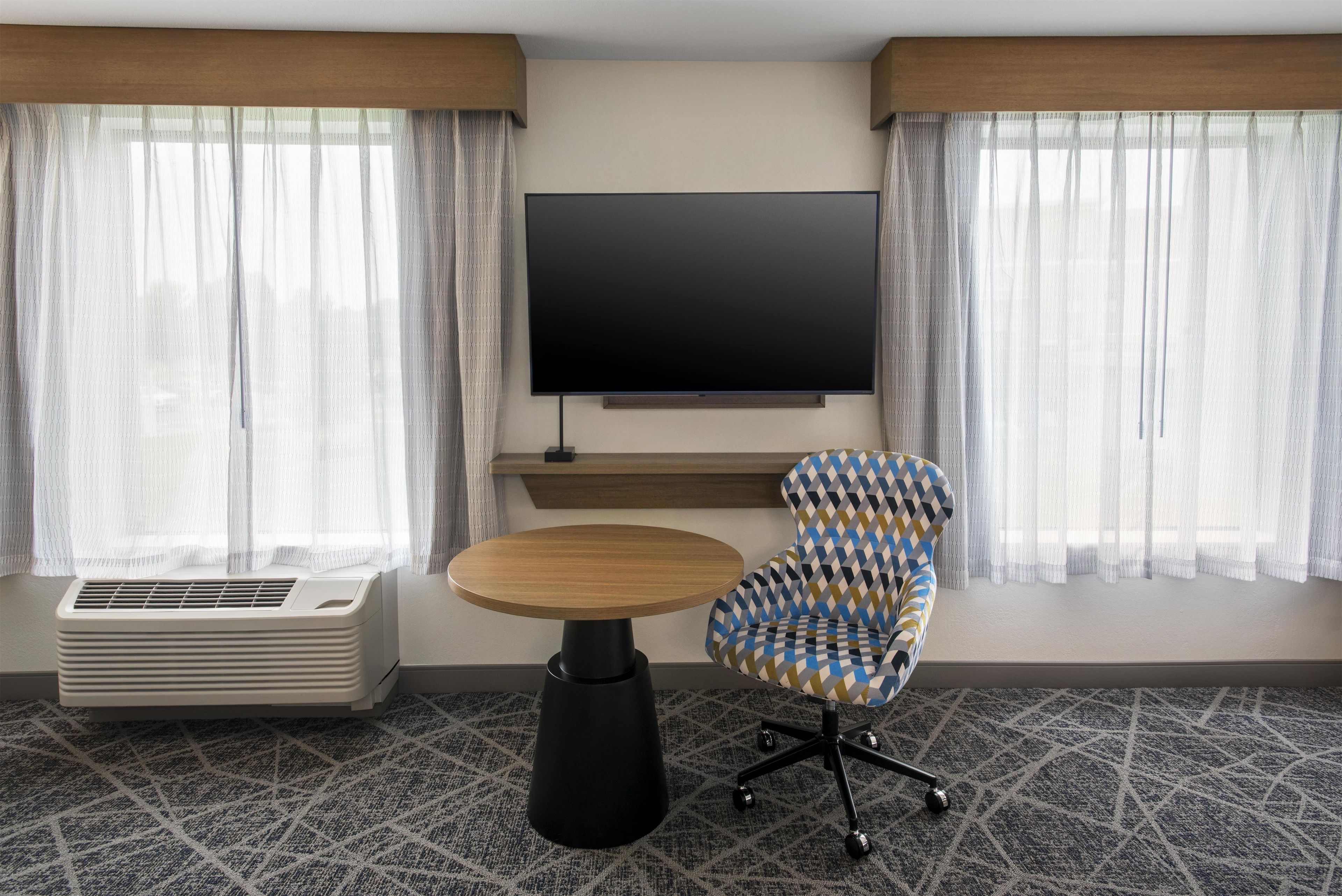 A hotel room with a flat-screen TV mounted on the wall, a round wooden table, a patterned armchair, and a portable air conditioner.