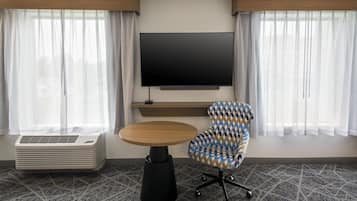 A hotel room with a flat-screen TV mounted on the wall, a round wooden table, a patterned armchair, and a portable air conditioner.