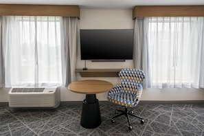 A hotel room with a flat-screen TV mounted on the wall, a round wooden table, a patterned armchair, and a portable air conditioner. - Hampton Inn and Suites New Hartford/Utica (New Hartford)