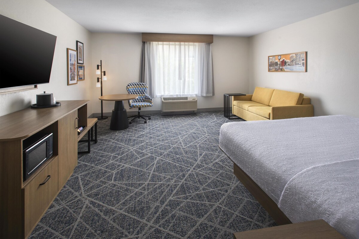 A hotel room with a bed, a sofa, a desk with a chair, a television, and a window with curtains.