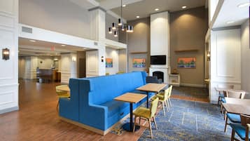 A hotel lobby with a blue sofa, wooden tables, and chairs. There are paintings on the walls and a TV mounted above a fireplace.