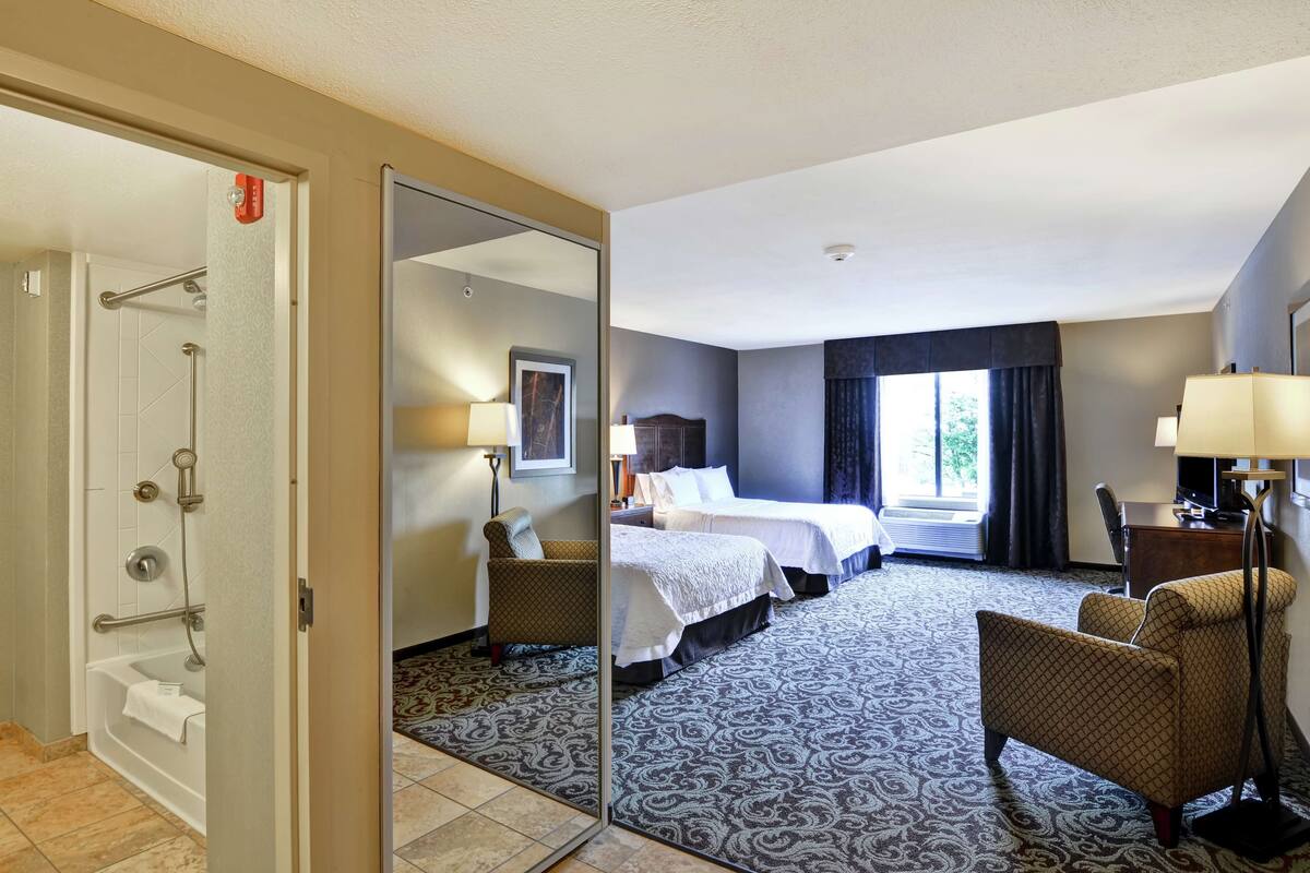 A hotel room with a large mirror, two beds, a desk, a chair, and a bathroom with a bathtub and shower.