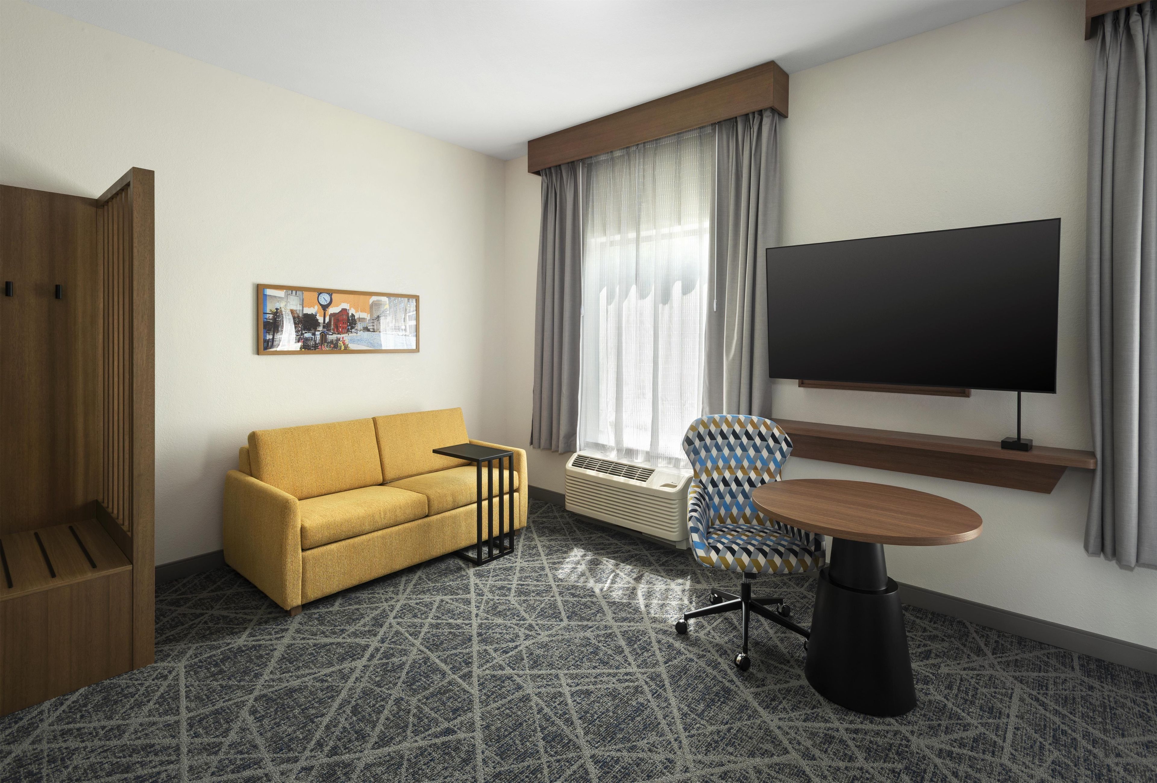 A hotel room with a sofa, a chair, a TV, a painting, and a window with curtains.