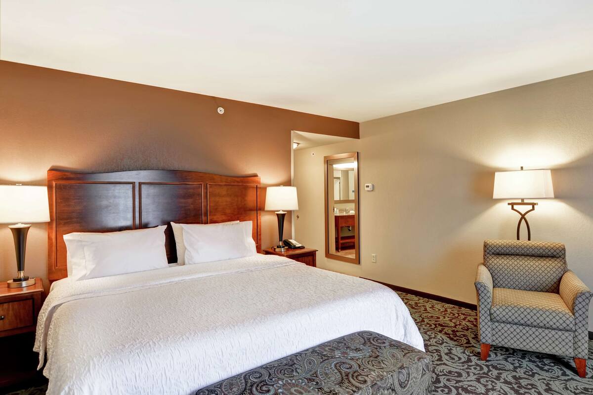 A hotel room with a large bed, two bedside tables with lamps, a chair, and a carpeted floor.
