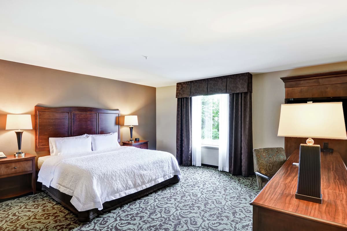 A hotel room with a large bed, two bedside tables with lamps, a desk with a lamp, a window with curtains, and patterned carpet.