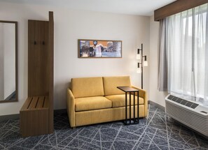 A hotel room with a sofa, a small table, a painting, a floor lamp, a mirror, and a window with sheer curtains. - Hampton Inn and Suites New Hartford/Utica (New Hartford)