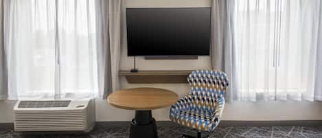 A hotel room with a flat-screen TV mounted on the wall, a round wooden table, a patterned armchair, and a portable air conditioner.