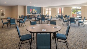 Une salle de conférence avec des tables rondes et des chaises bleues, un sol recouvert de moquette et de grandes fenêtres donnant sur un parking.