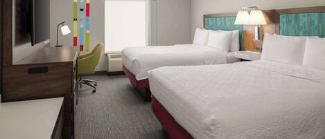 A hotel room with two beds, a desk, a TV, a desk lamp, and a window with blinds.