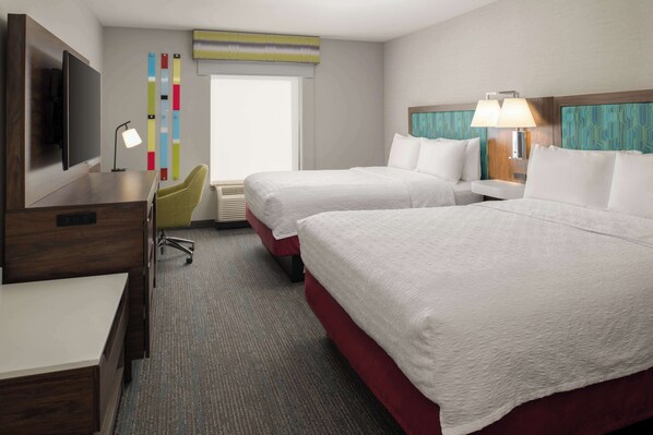 A hotel room with two beds, a desk, a TV, a desk lamp, and a window with blinds.