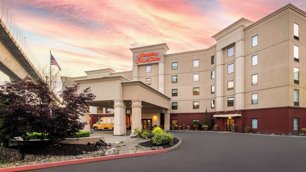 Un hôtel Hampton Inn & Suites au design moderne, avec une entrée imposante, un drapeau et des espaces verts bien entretenus.