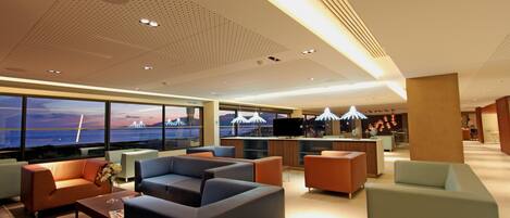 Um lobby moderno de hotel com áreas de estar, um balcão de recepção e grandes janelas que oferecem vista para uma ponte e o pôr do sol.