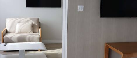A modern hotel room with a sofa, a small table, a TV mounted on the wall, and a wooden floor.