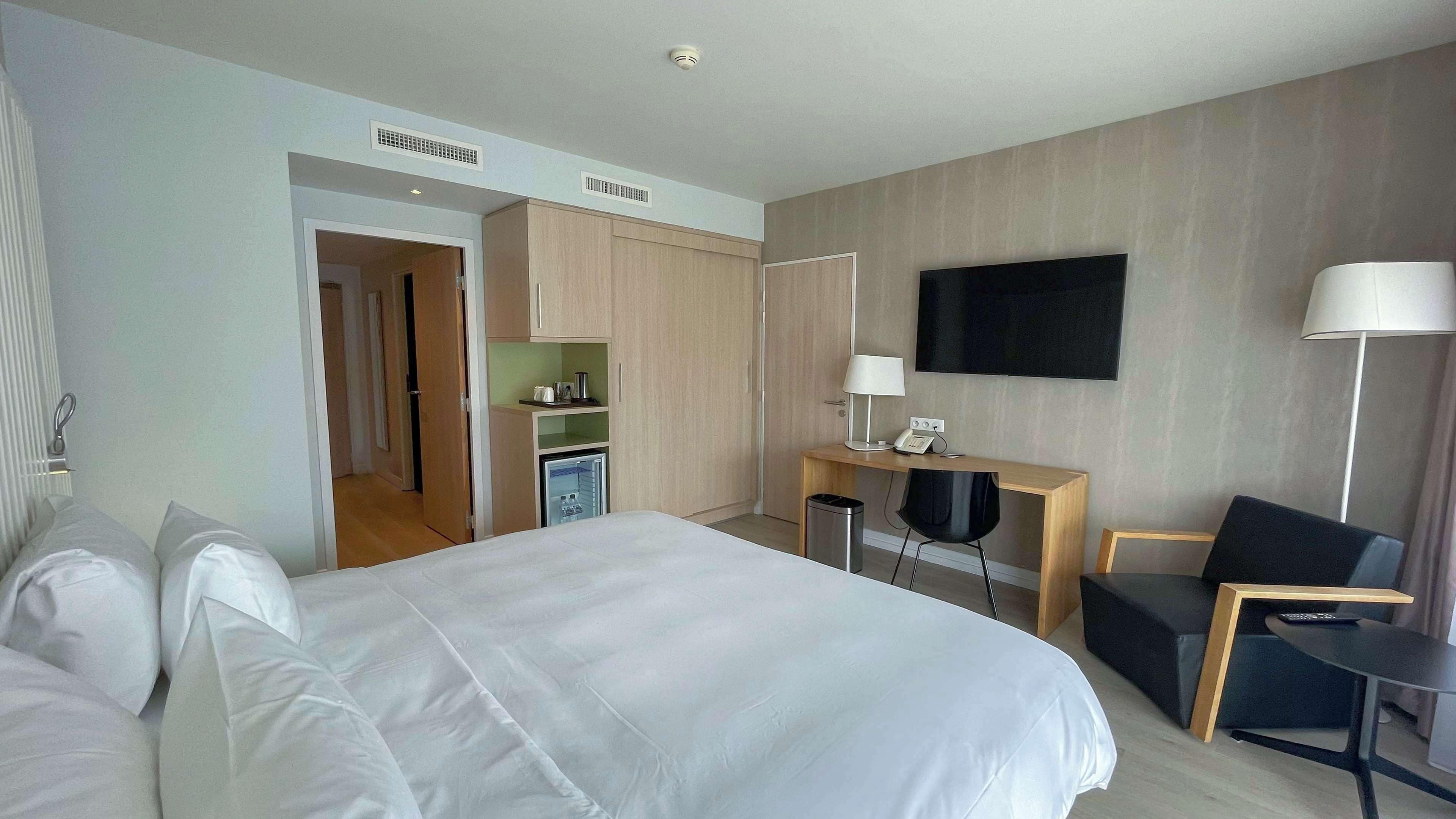 A hotel room with a large bed, a desk with a chair, a TV mounted on the wall, and a lamp.