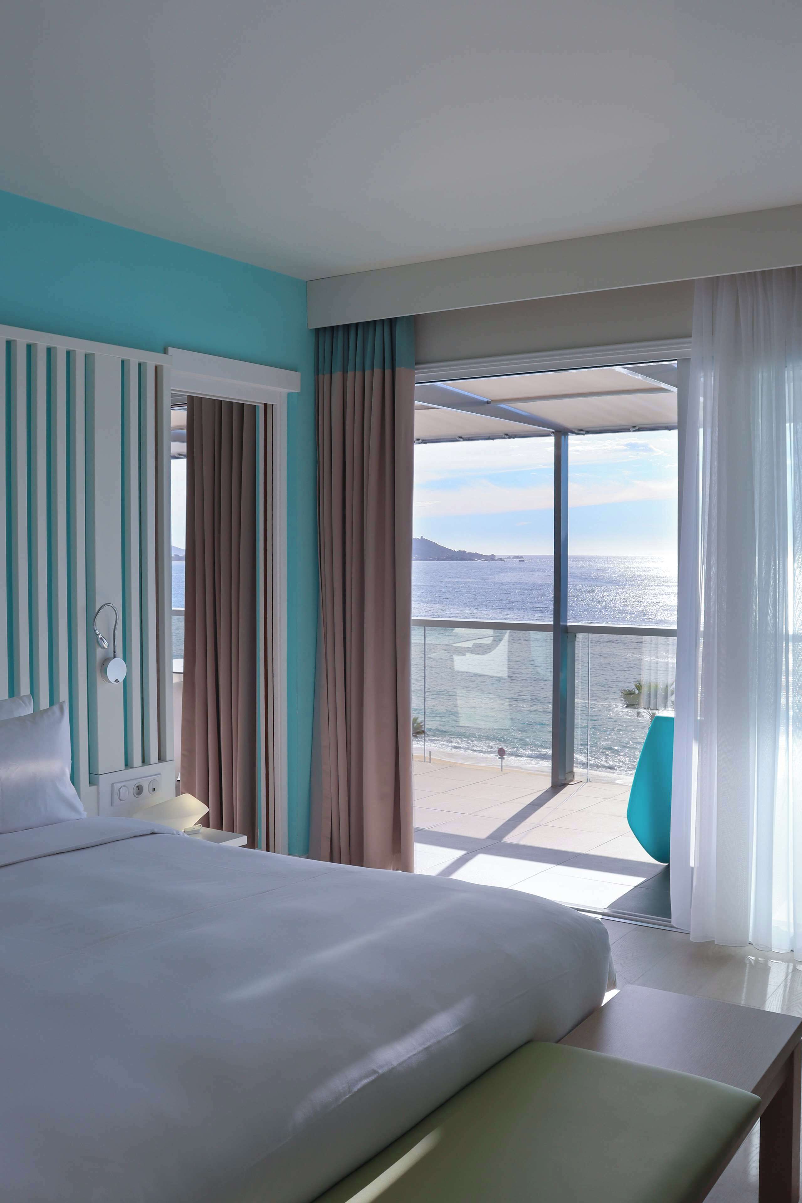 A hotel room with a bed, a bedside table, a window with curtains, and a view of the sea.