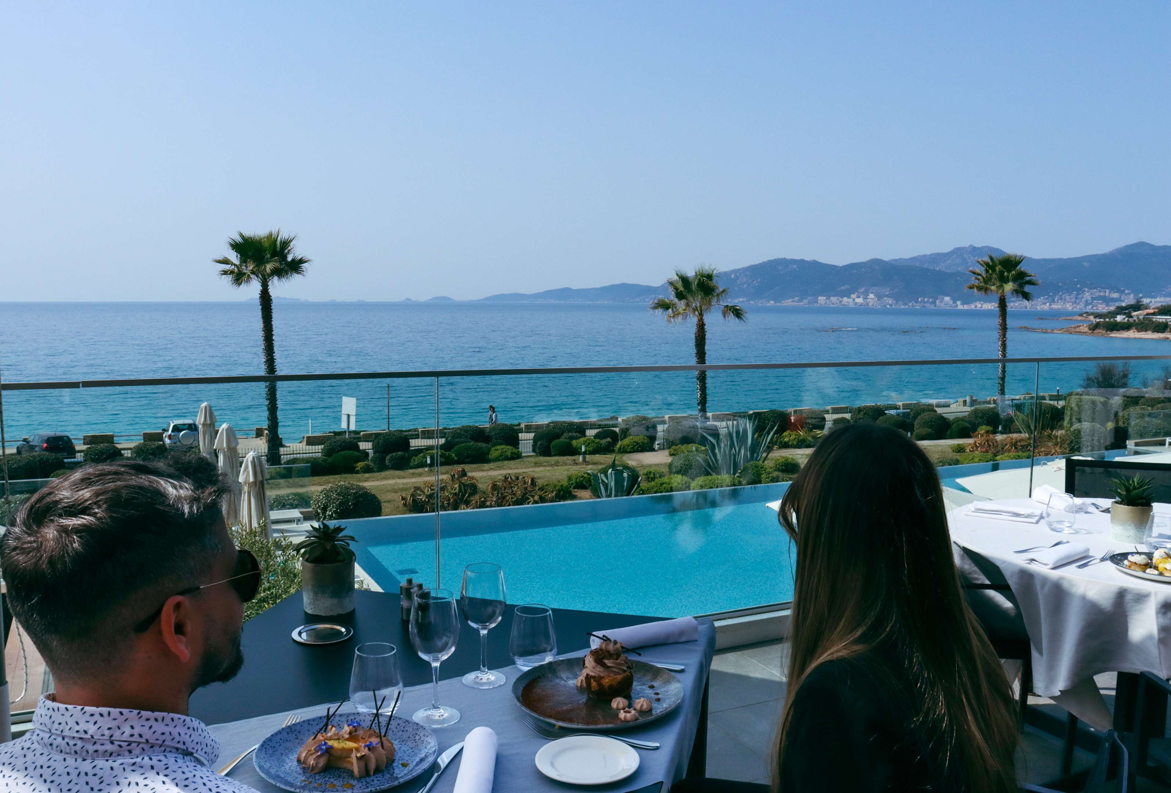 A dining table with a view of the ocean and mountains, a pool, and palm trees.