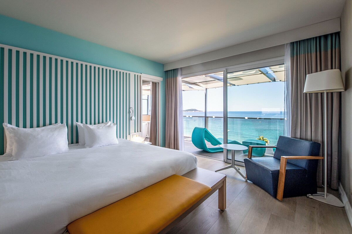 A hotel room with a bed, a bench, a chair, a small table, and a view of the sea.