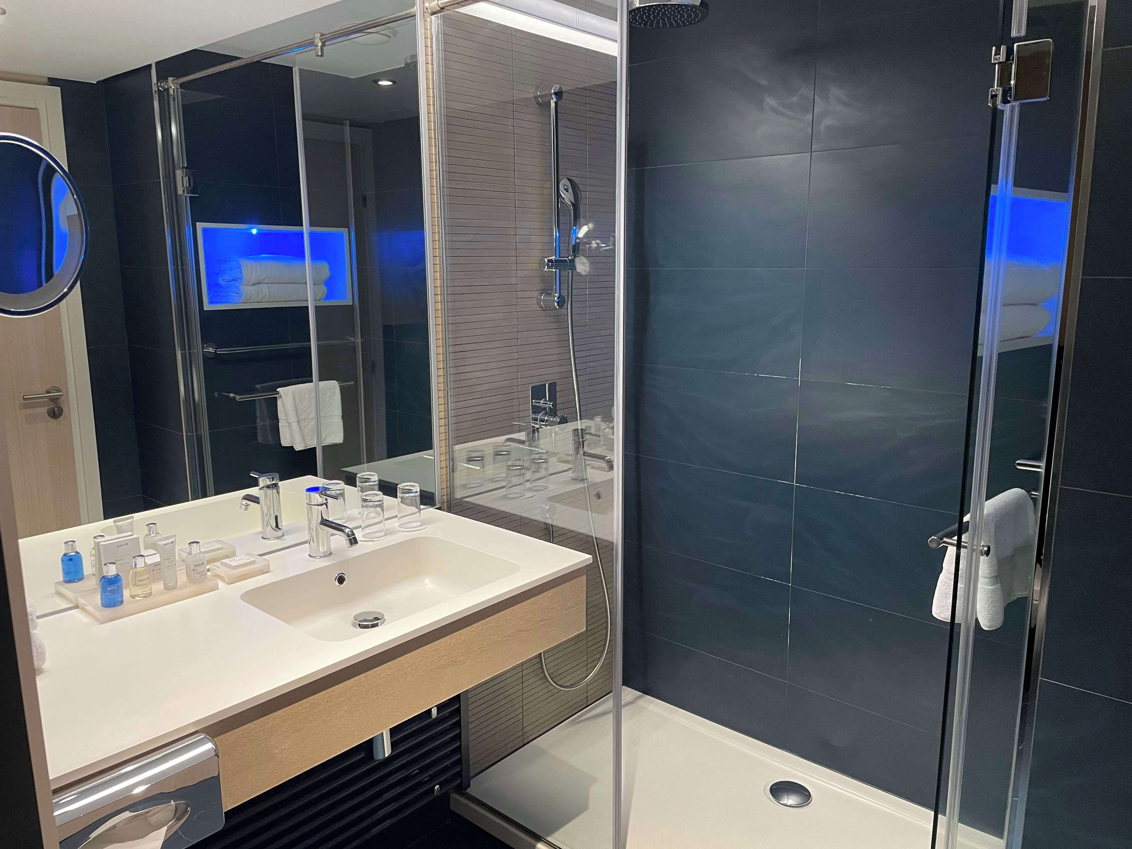 Un baño moderno con cabina de ducha de vidrio, un espejo grande y un tocador con múltiples artículos de aseo.