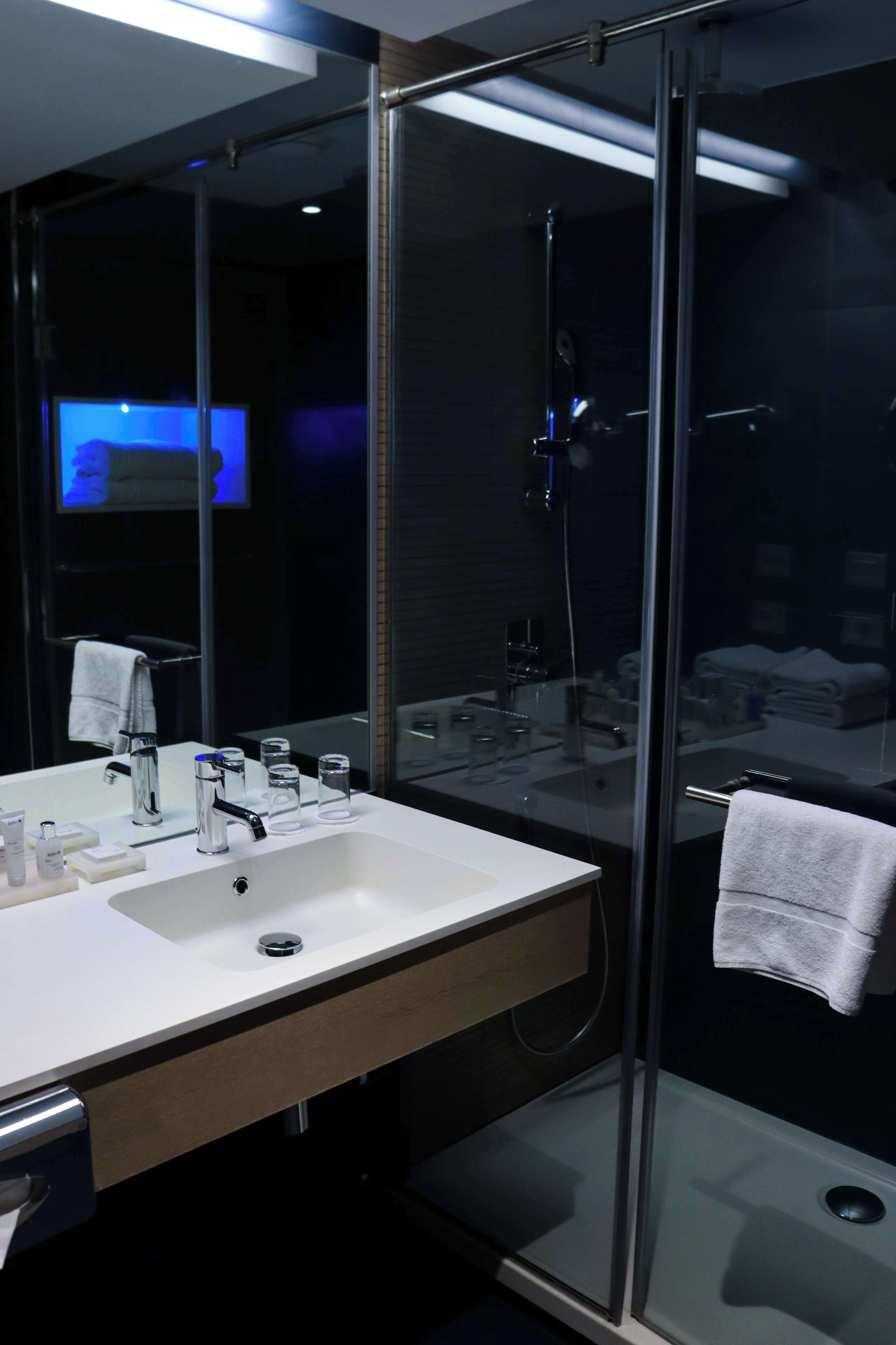 Un baño moderno con mampara de vidrio, un espejo grande y un lavamanos blanco con artículos de aseo.