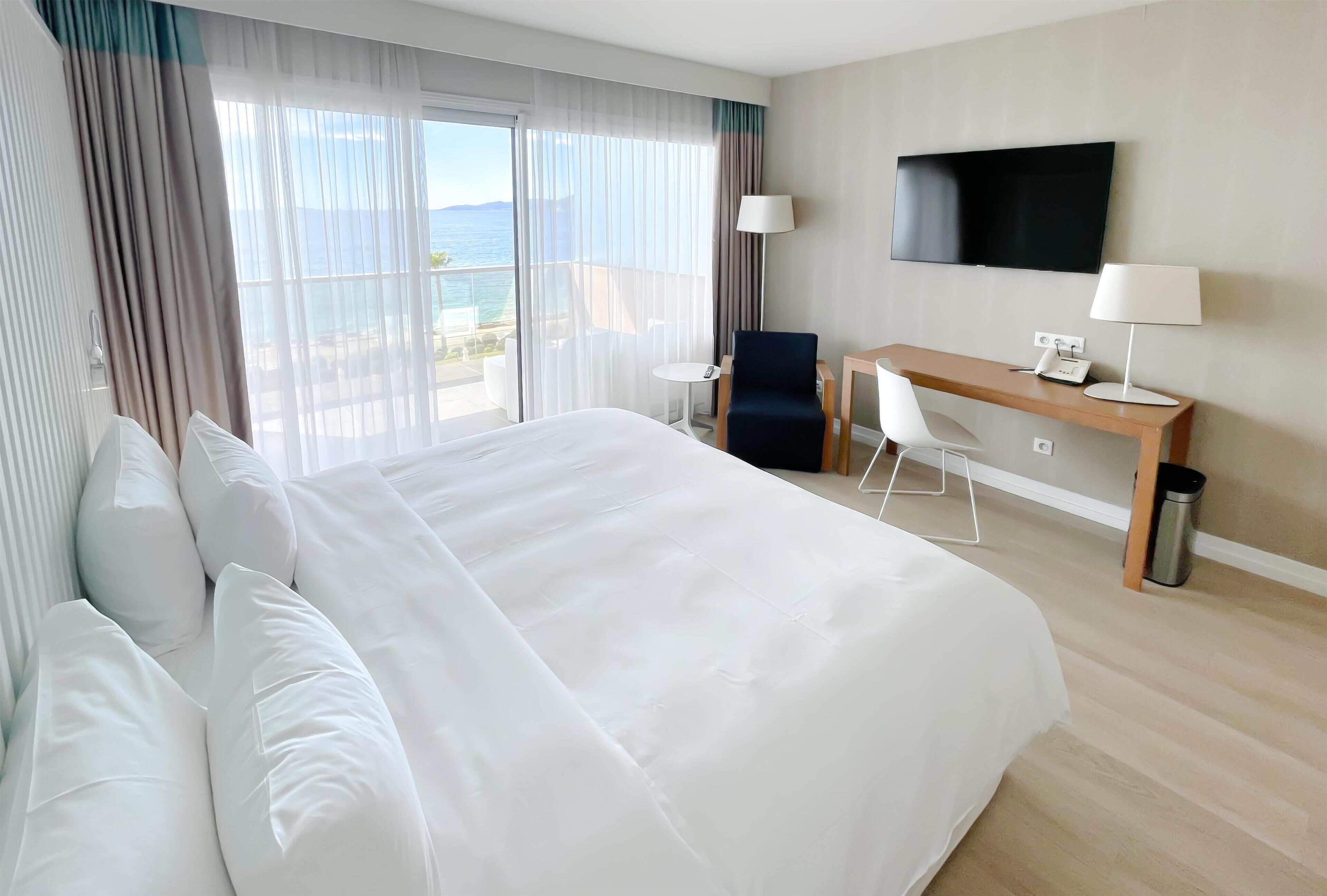 A hotel room with a large bed, a desk, a chair, a TV, and a view of the sea.