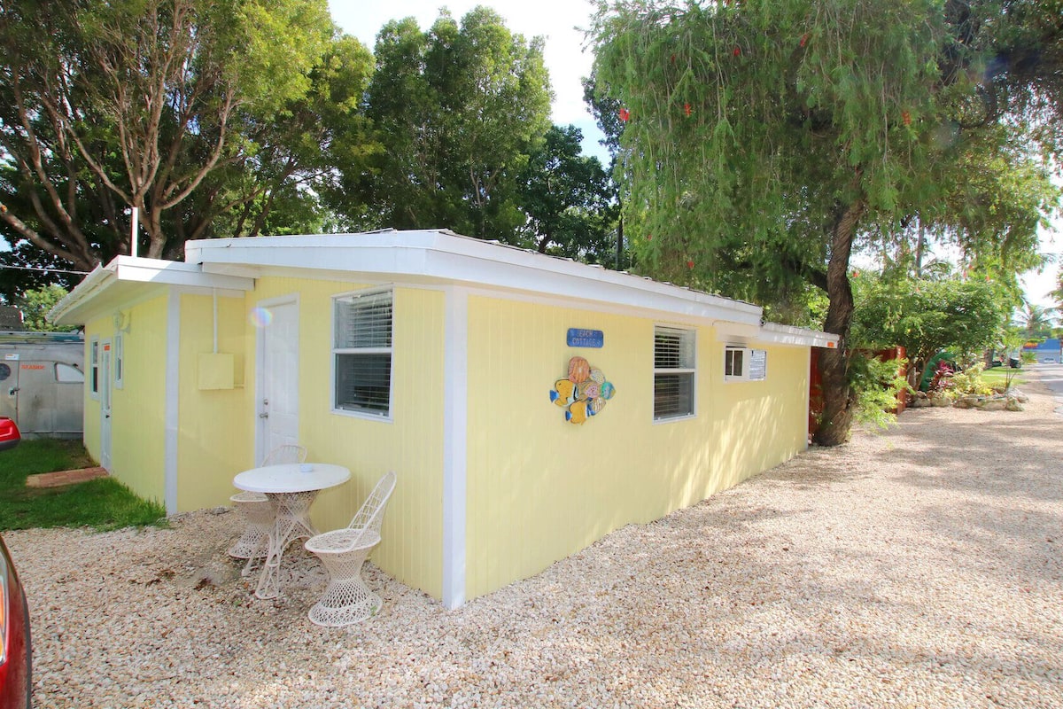 A small, yellow cottage-style building with a white door and windows, a small table with chairs, and a sign indicating it's a cottage.