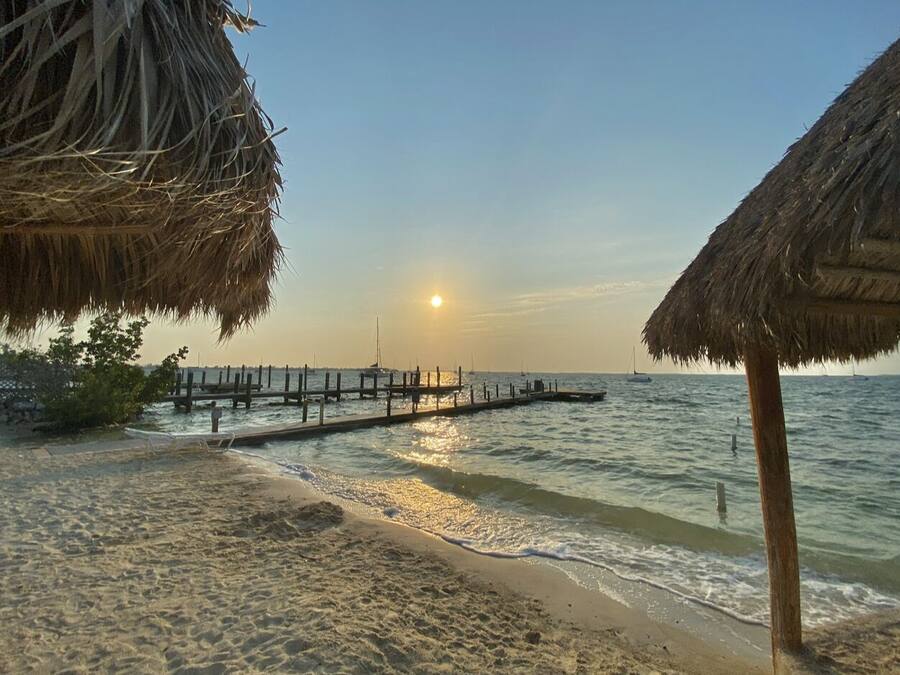 A beach with a wooden pier extending into calm waters, a thatched-roof structure on the left, and another on the right.