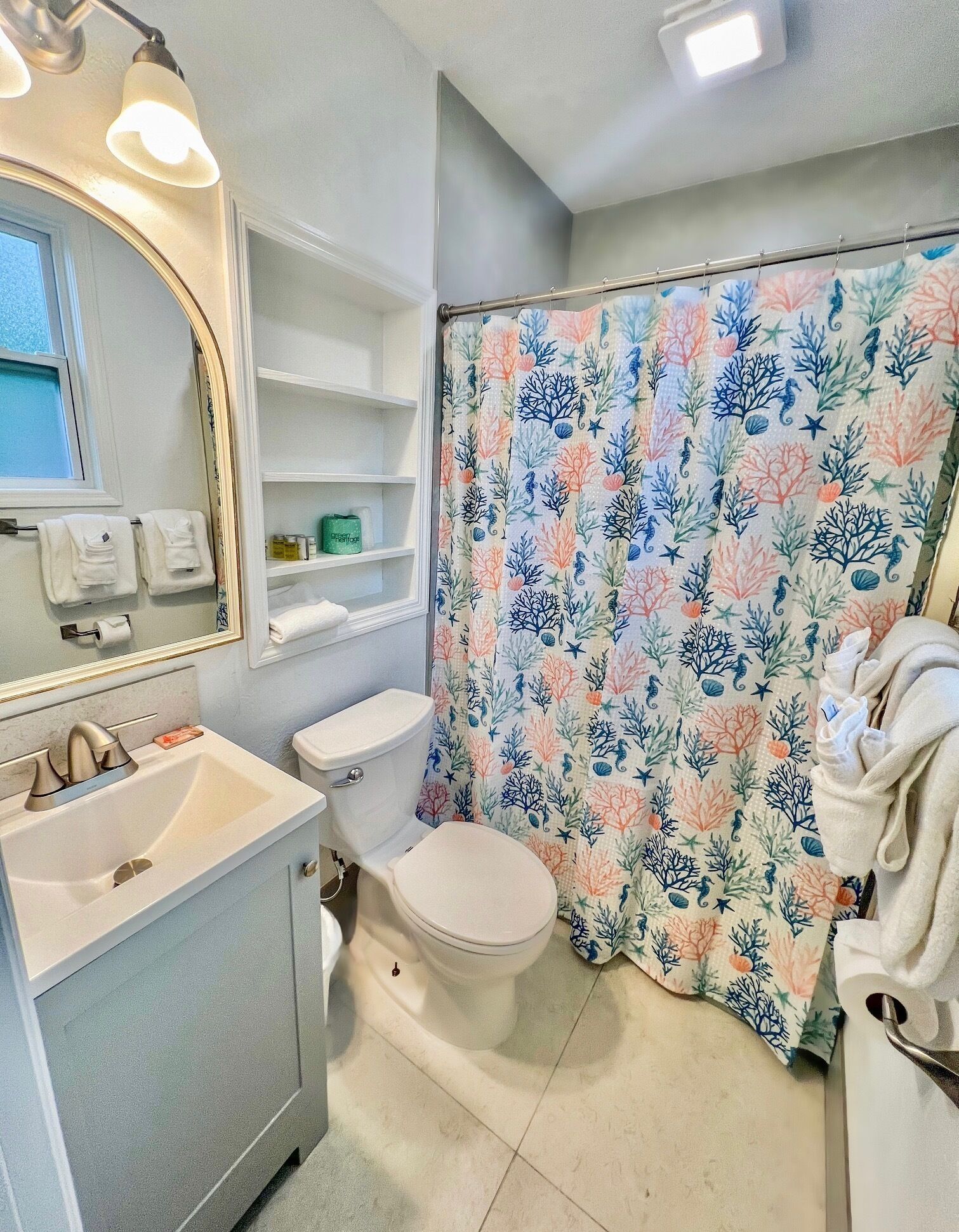 Un baño con un lavamanos blanco, un inodoro, una cortina de ducha con estampado floral y un espejo con luces arriba.