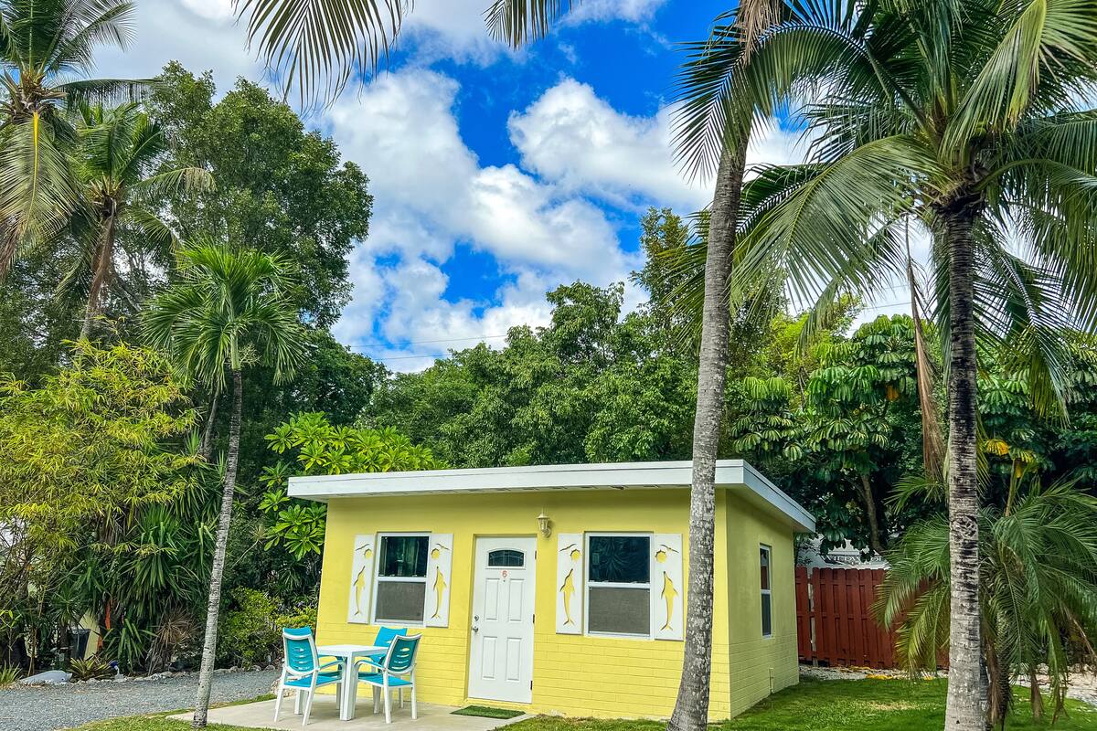 A small yellow building with a white door and windows, surrounded by palm trees and greenery.
