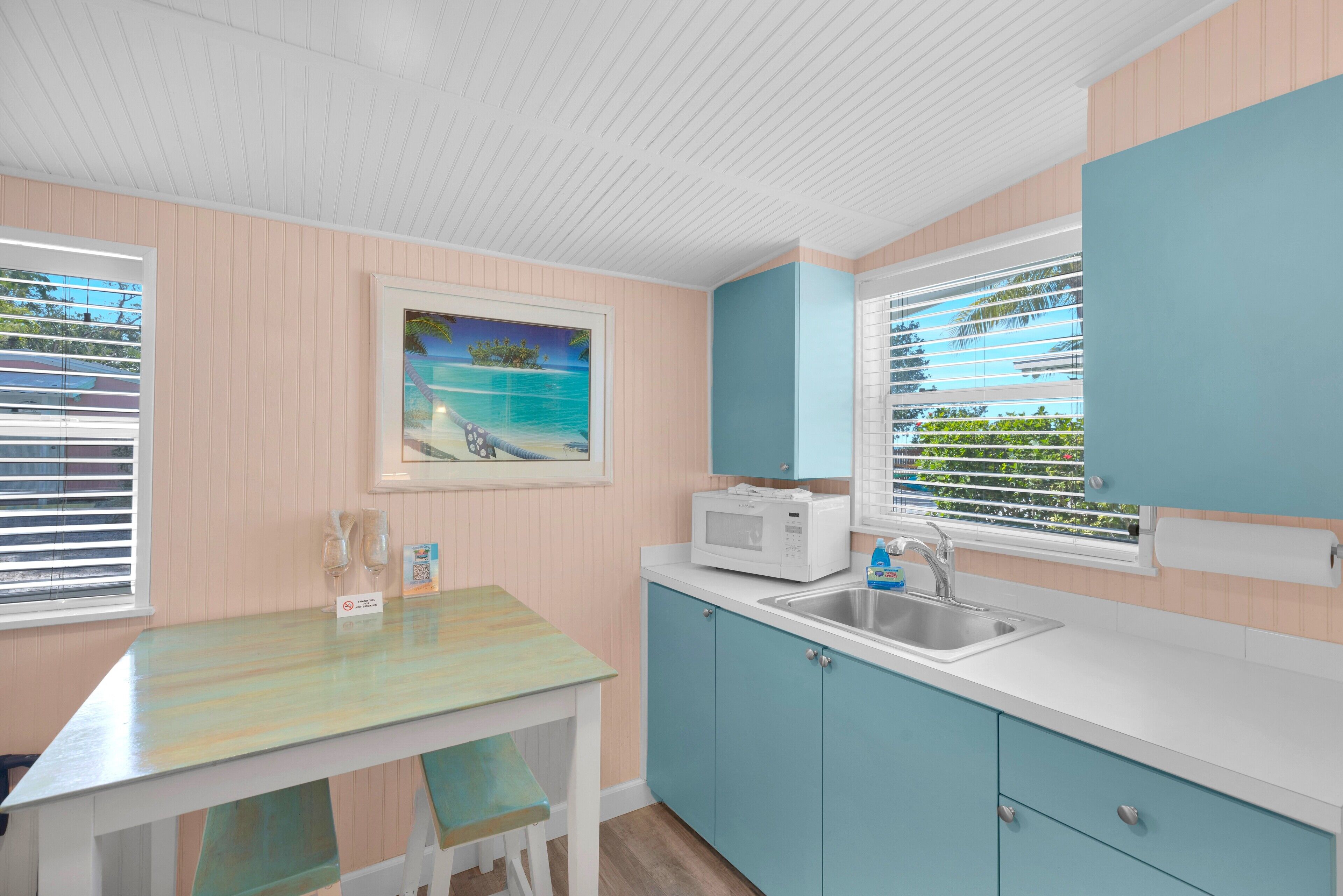 A kitchen with a white countertop, blue cabinets, a microwave, and a sink. There is a painting of a beach scene on the wall.