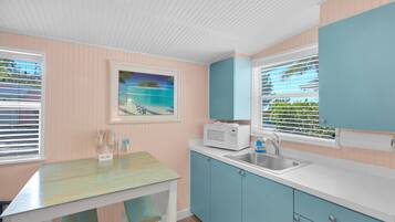A kitchen with a white countertop, blue cabinets, a microwave, and a sink. There is a painting of a beach scene on the wall.