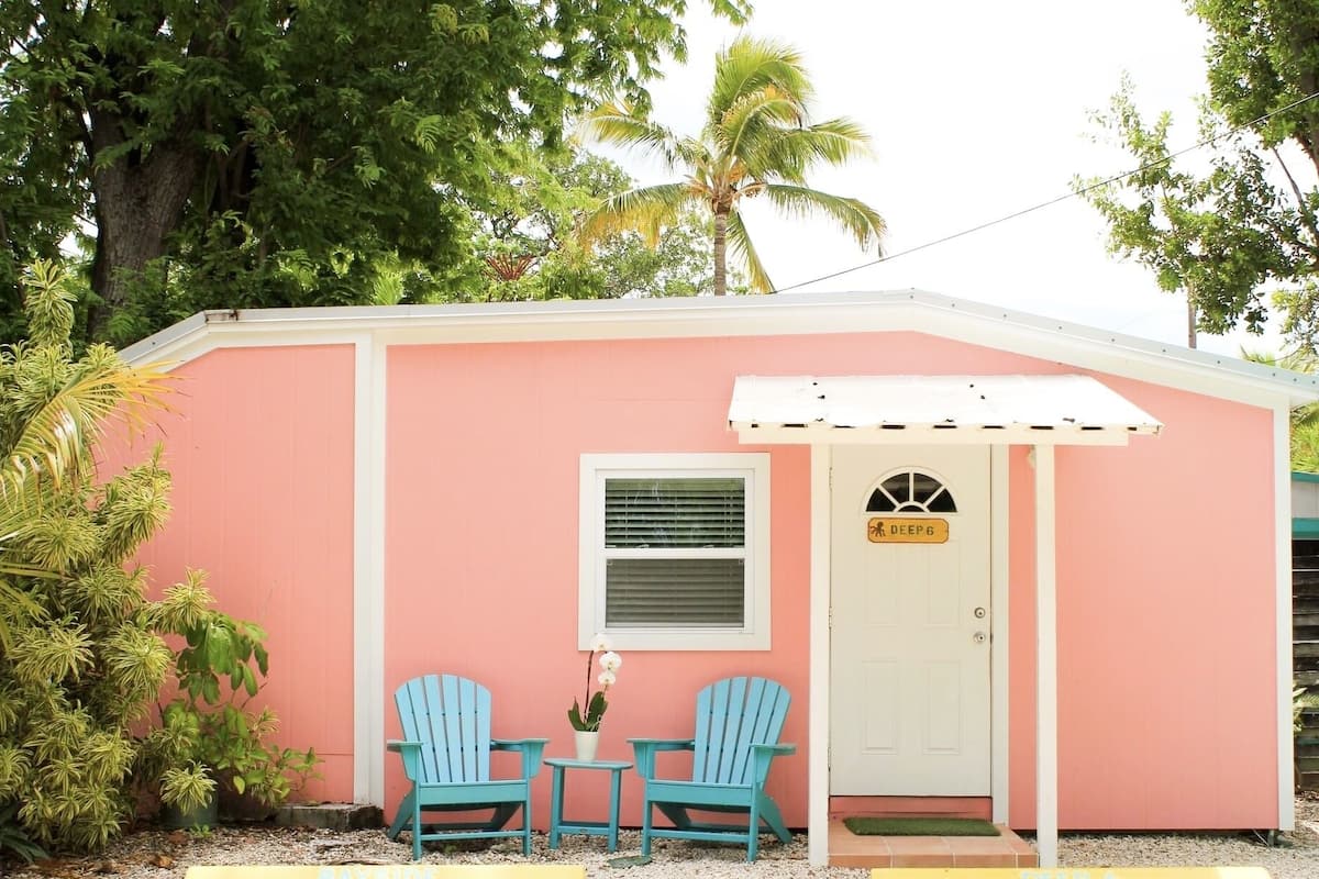 A small pink building with a white door labeled "DEEP 6". There are two blue Adirondack chairs and a small table with a potted plant in front.