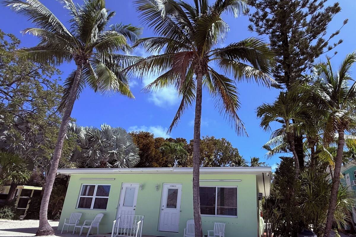 A small, single-story building with a light green exterior, white-framed windows, and a door. There are white plastic chairs outside, and palm trees are visible in the background.